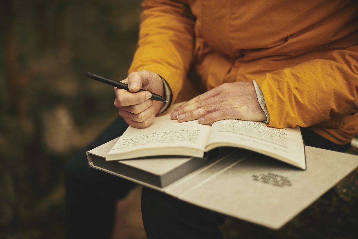 person holding a journal, pen and a book in their lap