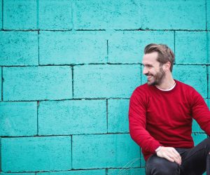 man in red shirt leaning against a teal wall